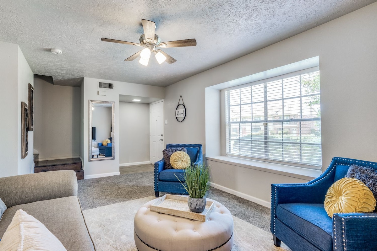 A bright living area is furnished with a comfortable sofa and two blue armchairs, complemented by a round ottoman. Natural light enters through a large window adorned with blinds, while a ceiling fan adds a functional touch to the space.
