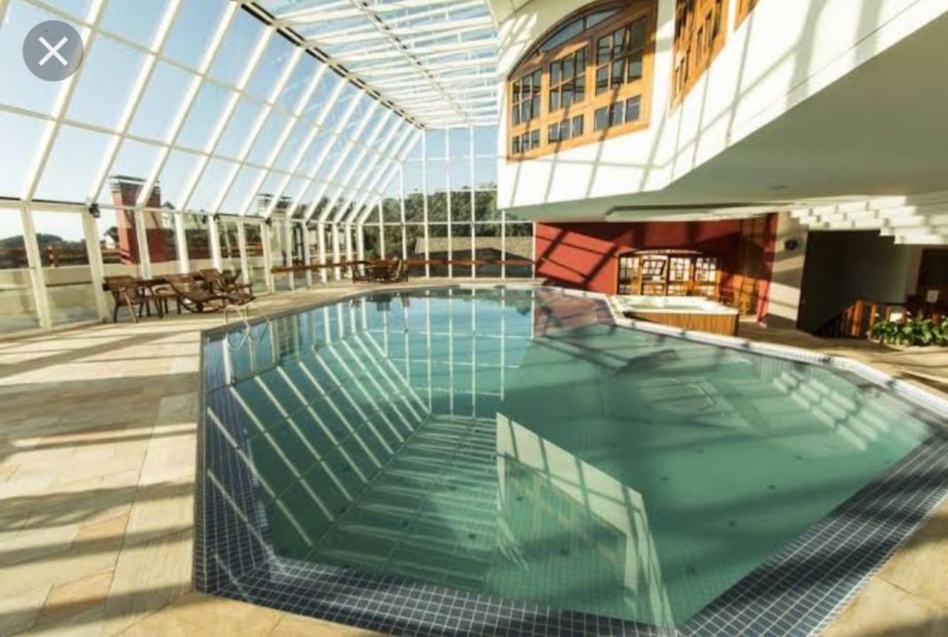 An indoor heated pool is surrounded by large glass panels, allowing natural light to illuminate the space. Reflective tiles create a serene atmosphere as sunlight casts patterns on the water's surface. Comfortable seating is arranged nearby, adding to the inviting environment.