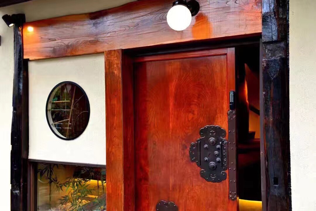 The entrance of the house features a beautifully crafted wooden door with intricate metal hardware. A circular window alongside adds an elegant touch, allowing natural light to filter in. Warm lighting enhances the welcoming atmosphere of the entryway.