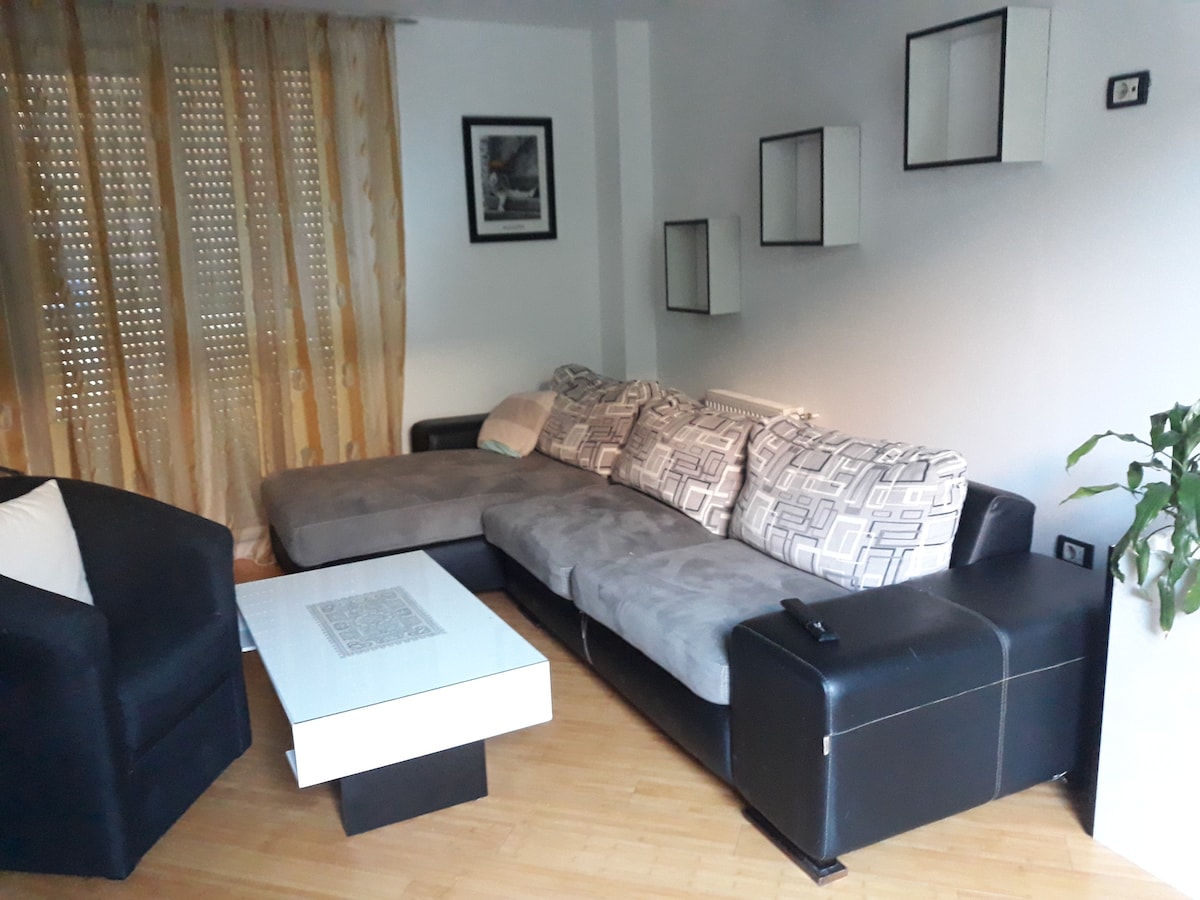 A spacious living area features a large sectional sofa with patterned upholstery, complemented by a modern coffee table. Decorative wall shelves enhance the minimalist aesthetic. Light filters through sheer curtains, creating a warm ambiance, while a potted plant adds a touch of greenery.