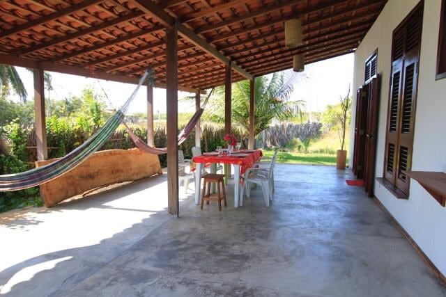 A spacious covered patio features a dining table set for meals, with plastic chairs and a wooden stool. Colorful hammocks hang nearby, surrounded by lush greenery. Natural light enters from the open sides, creating a relaxing outdoor atmosphere.