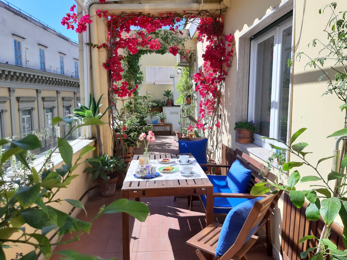 A sunny terrace features a wooden dining table set with dishware and food. Blue chairs surround the table, complemented by vibrant pink bougainvillea and green plants. The backdrop includes classic architecture and additional greenery, creating a tranquil outdoor dining space.