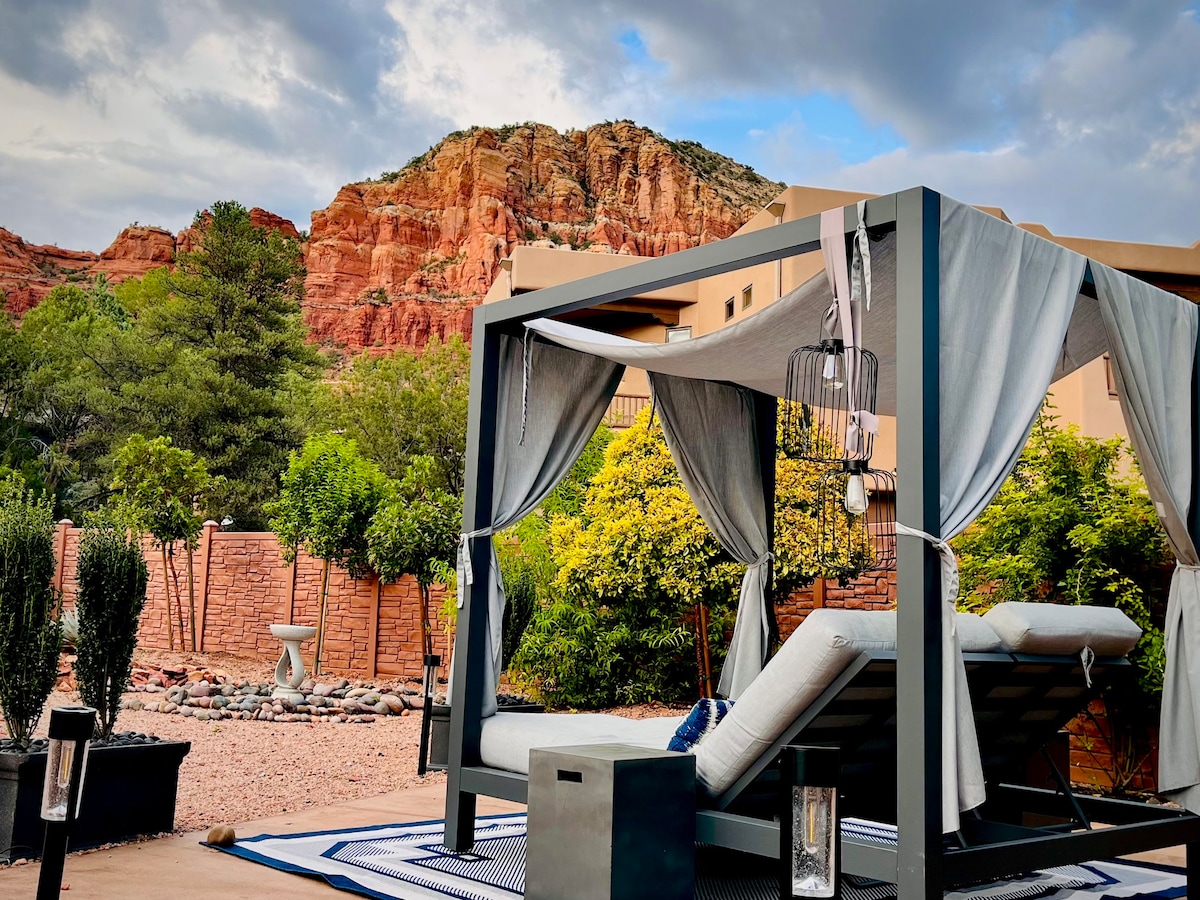 A private outdoor cabana featuring plush lounge beds is positioned against a backdrop of Red Rock formations. Greenery and desert landscaping surround the space, while lanterns add a gentle ambiance. The sky is filled with soft clouds, highlighting the relaxed atmosphere.
