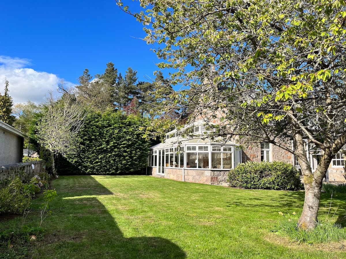 A spacious garden area features well-maintained grass surrounded by trees and shrubs. A large conservatory is seen in the background, with sunlight illuminating the scene under a clear blue sky.