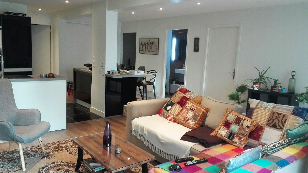 A spacious living area features a light-colored sofa adorned with colorful cushions, accompanied by a sleek wooden coffee table. A stylish armchair and decorative rug contribute to the cozy atmosphere. The open layout connects to a modern kitchen, visible in the background.