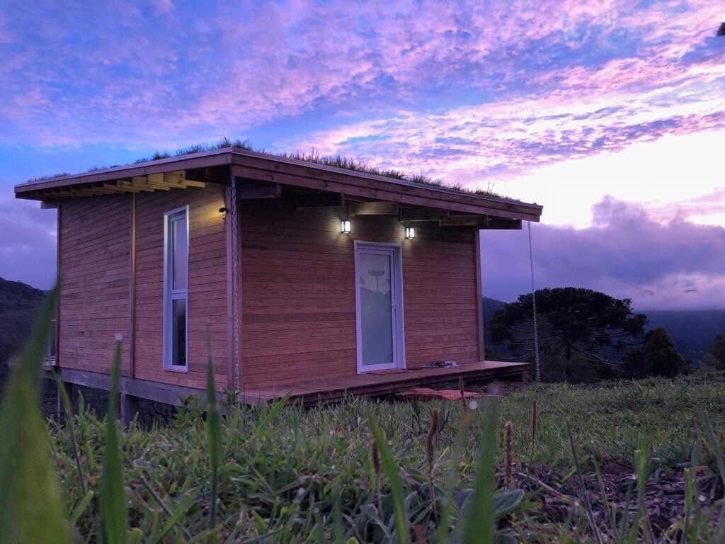 A wooden cabin perched on a grassy hilltop features a green roof and large windows. Soft evening light enhances the warm tones of the wood, while a colorful sky provides a scenic backdrop. The surrounding landscape features lush greenery and distant hills.