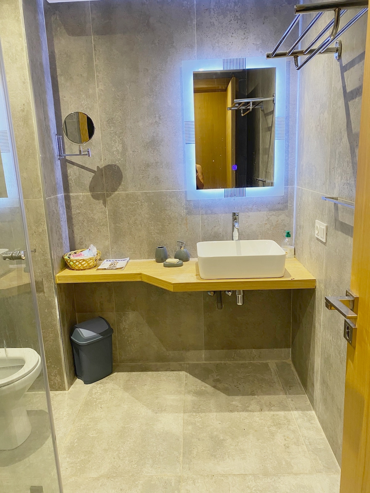 A modern bathroom features sleek gray tiles and a spacious walk-in shower with a glass divider. A basin with clean lines is placed on a wooden countertop, complemented by a circular mirror with integrated lighting, providing a bright and functional space.