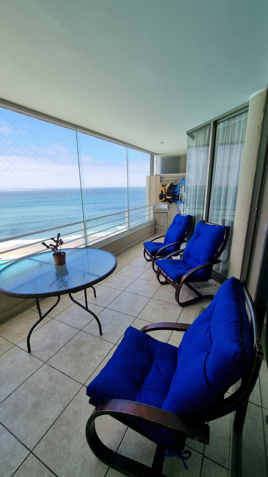 A closed terrace features two blue lounge chairs and a round glass table, creating a space to relax. The stunning view of the ocean is visible through large glass windows, with the tranquil sea in the background. Tiled flooring contributes to the modern ambiance.