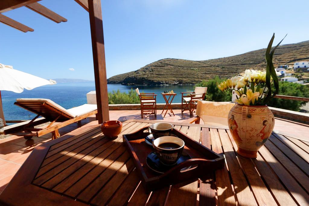 An outdoor dining area is visible, featuring a wooden table with a tray holding two cups of coffee. A vase of flowers sits on the table, while wooden chairs and a sunbed are arranged nearby. The tranquil coastline and hills can be seen in the background under a clear blue sky.