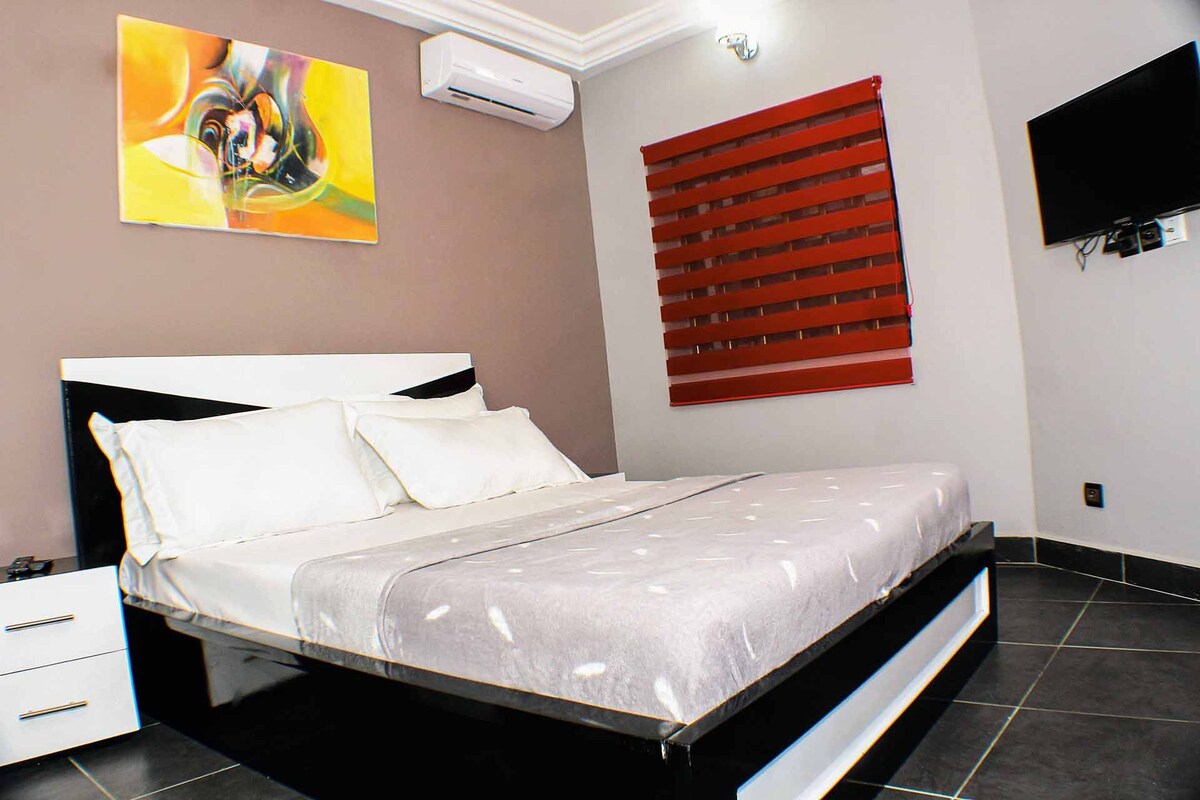 A spacious sleeping area features a large bed with crisp white linens and a light gray blanket. The room is decorated with a colorful abstract painting and equipped with a flat-screen TV mounted on the wall. Air conditioning and red window blinds add functionality and style.