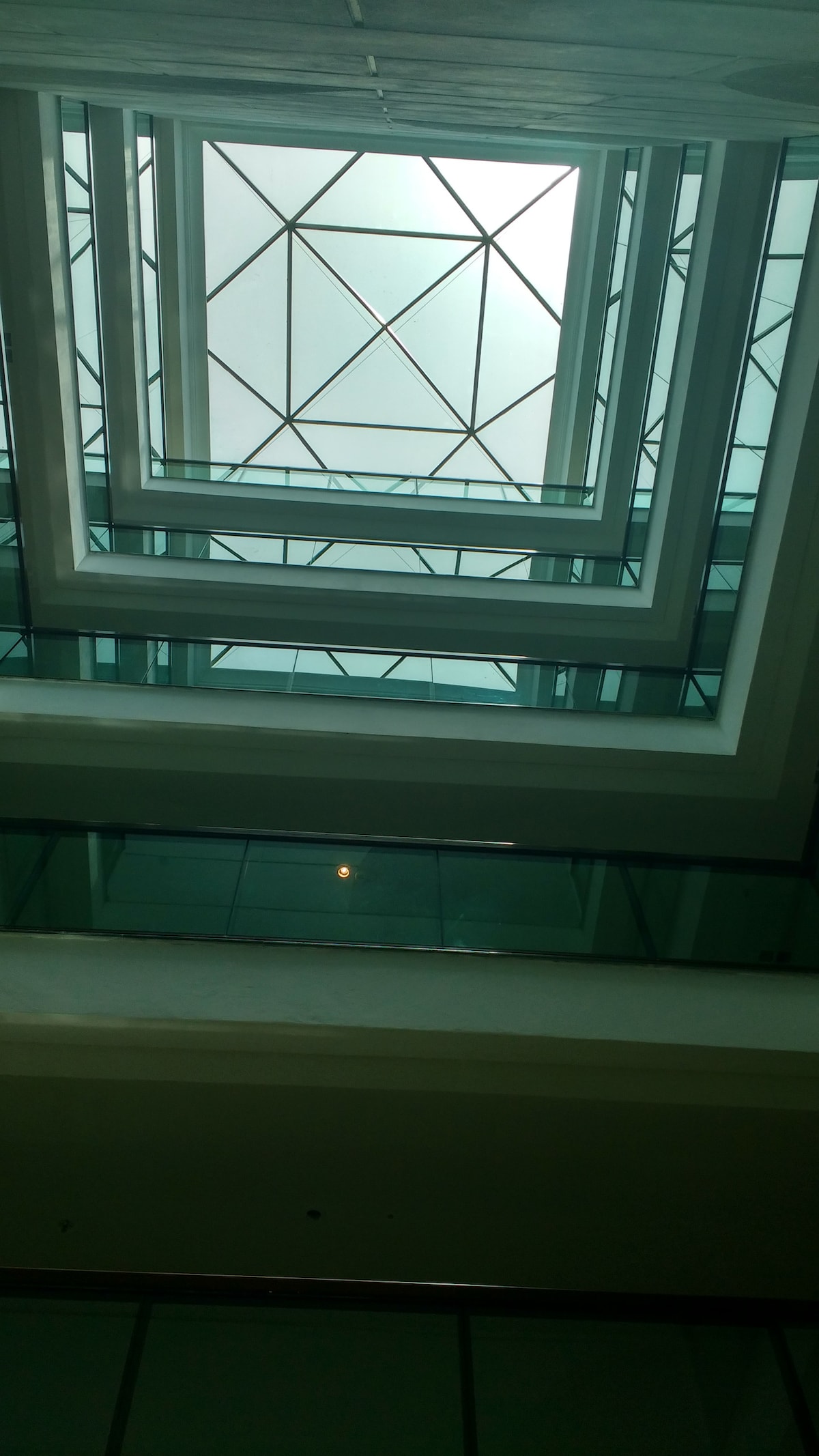 A geometric glass skylight allows natural light to illuminate the interior space, creating an intricate visual effect through multiple levels. The design incorporates triangular and rectangular shapes, with a clear view of the blue sky above, enhancing the sense of openness and connection to the outdoors.