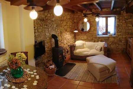 A cozy living area features a stone wall and wooden beams. A white sofa with decorative pillows is situated beside a small table adorned with flowers. A wood-burning stove provides warmth, while large windows allow natural light to fill the space.