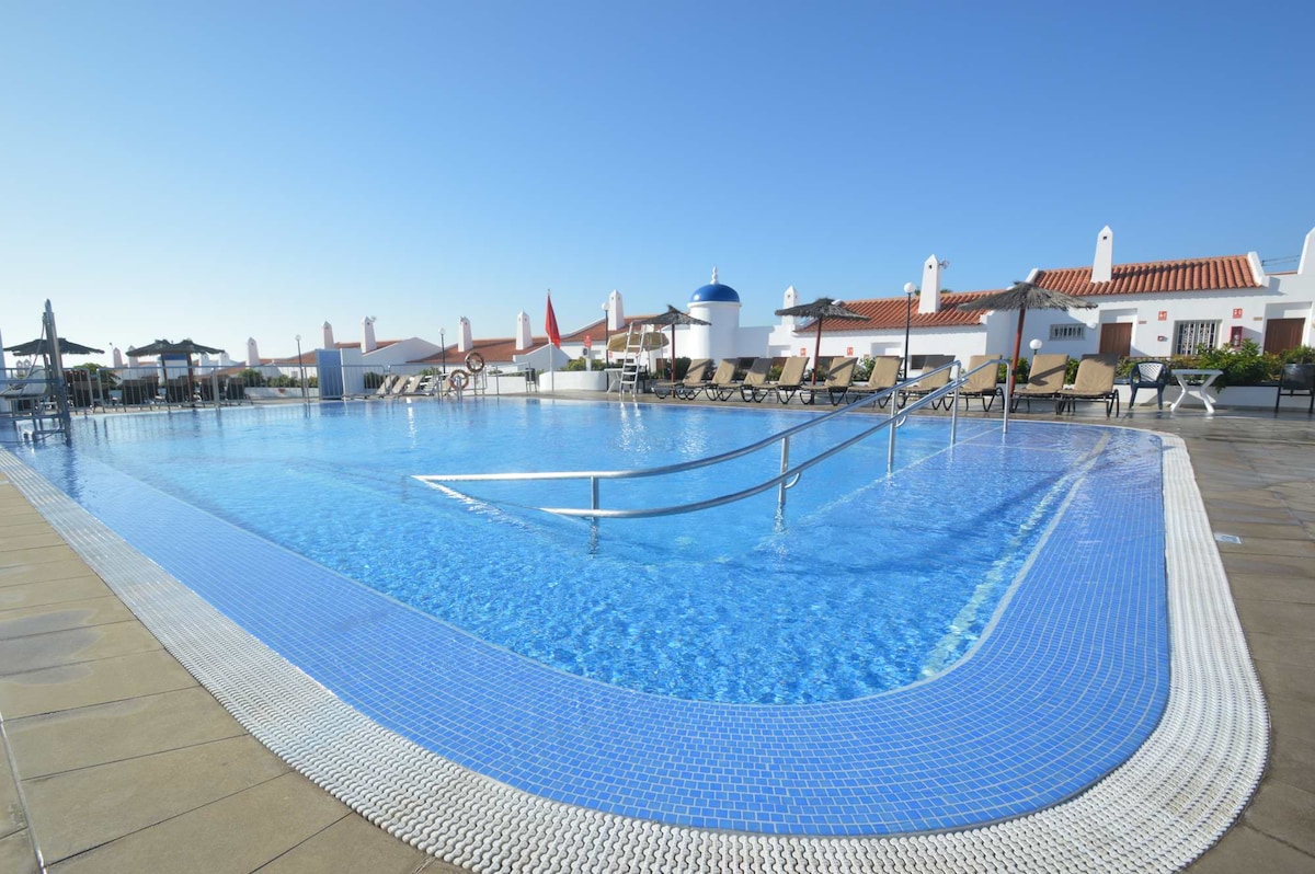 A spacious pool area is depicted, featuring a large, shimmering blue pool with a gradual entry and a textured edge. Surrounding loungers and shaded umbrellas provide ample seating. The backdrop includes whitewashed buildings with red roofs, under a clear blue sky.