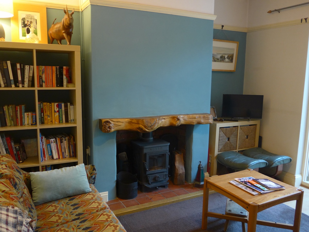 A snug living area is featured with a comfortable sofa, a wooden coffee table, and a small television. A wood burner is positioned within a brick fireplace, complemented by a rustic wooden mantel. A bookshelf displays various books, adding character to the space.