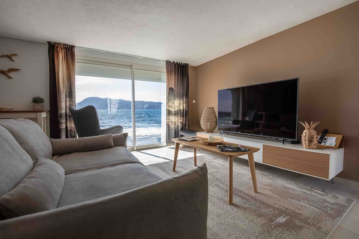 A cozy living area features a light-colored sofa and a wooden coffee table. A large television sits on a sleek media console. Floor-to-ceiling windows offer direct views of the beach and surrounding landscape, allowing natural light to fill the space.