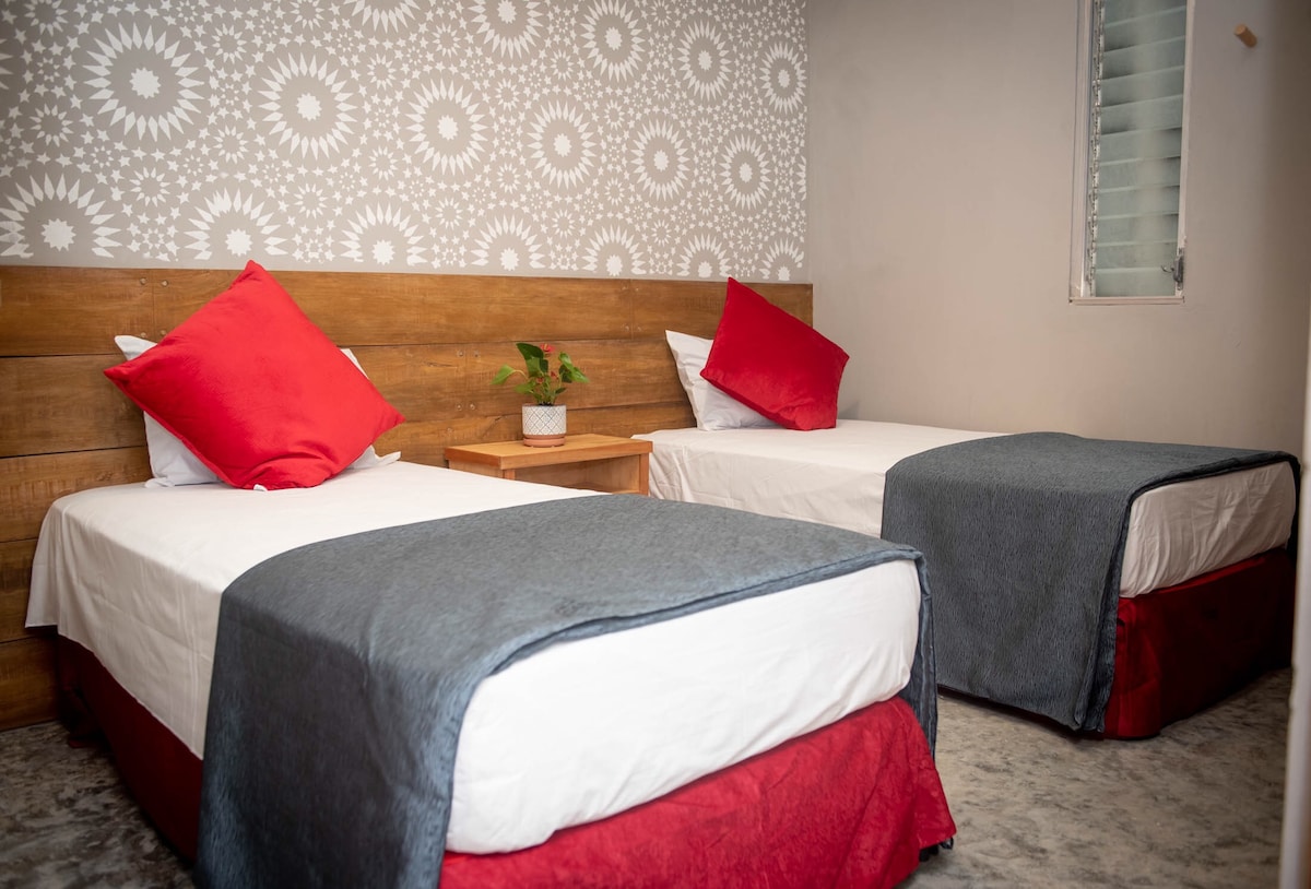A spacious room is displayed, featuring two twin beds with red-accented bed skirts and grey blankets. Each bed is accompanied by a wooden nightstand. A decorative plant sits on the nightstand, and a patterned wall adds character to the space. A window allows natural light to enter.