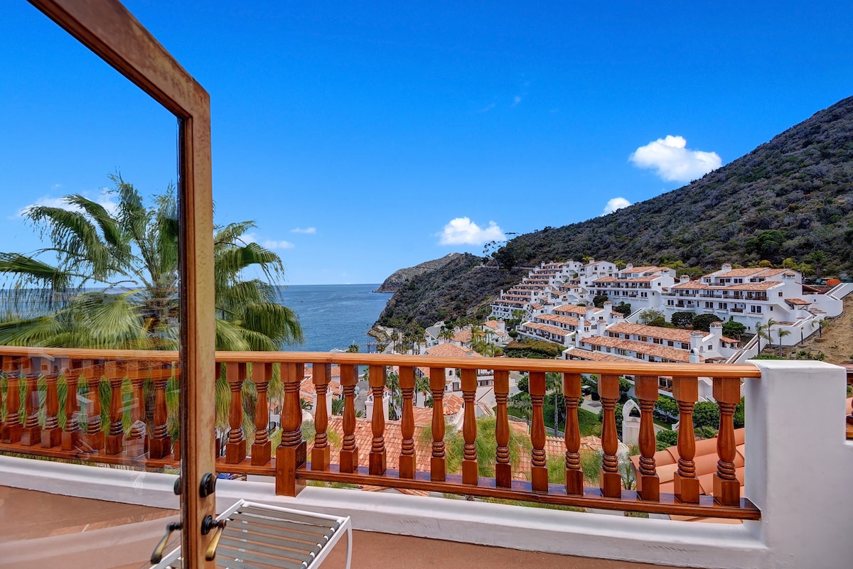 A spacious wrap-around balcony frames ocean views and nearby hillside residences. Lush greenery and bright blue sky create a serene setting. The railing features decorative wooden slats, inviting outdoor relaxation and enjoyment of the expansive scenery.