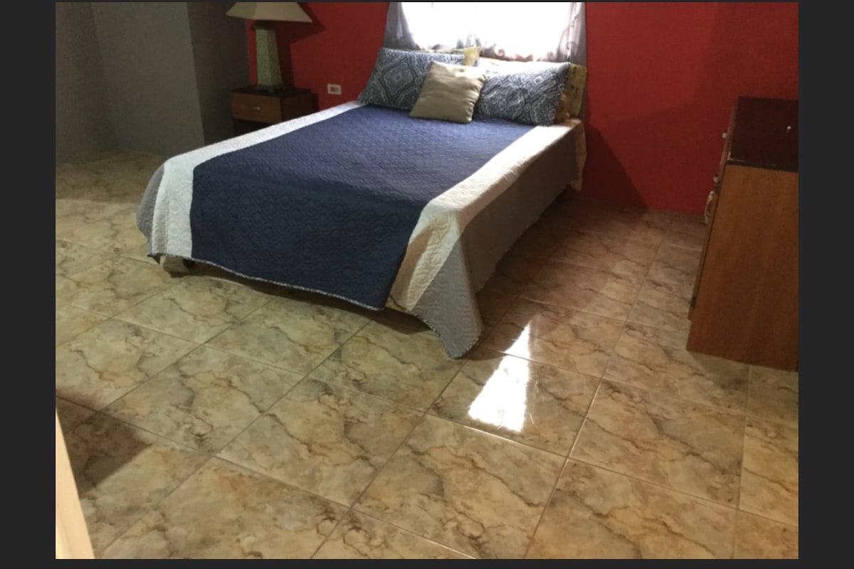 A spacious bedroom is shown, featuring a large bed covered with a patterned blanket. A bedside lamp is present on a wooden nightstand, and a dresser occupies one corner. Natural light enters through a window, illuminating the tiled floor.