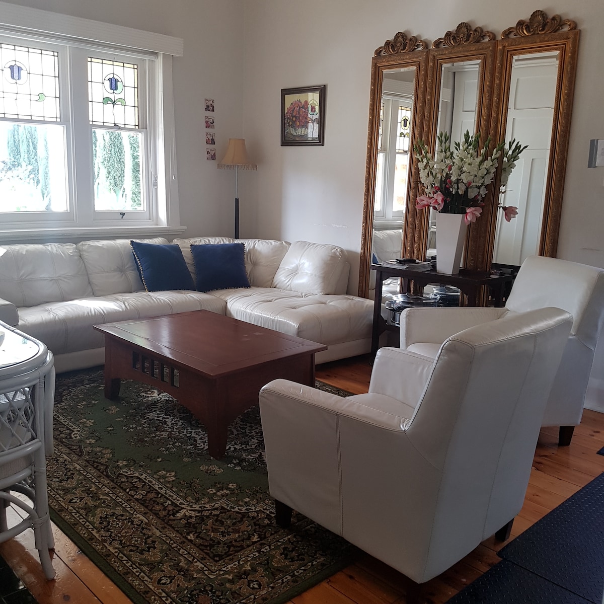 A bright living room features a large white sectional sofa and two white armchairs. A wooden coffee table is positioned centrally on an area rug, complemented by a decorative mirror and floral arrangement. Natural light filters through a window adorned with colorful stained glass.
