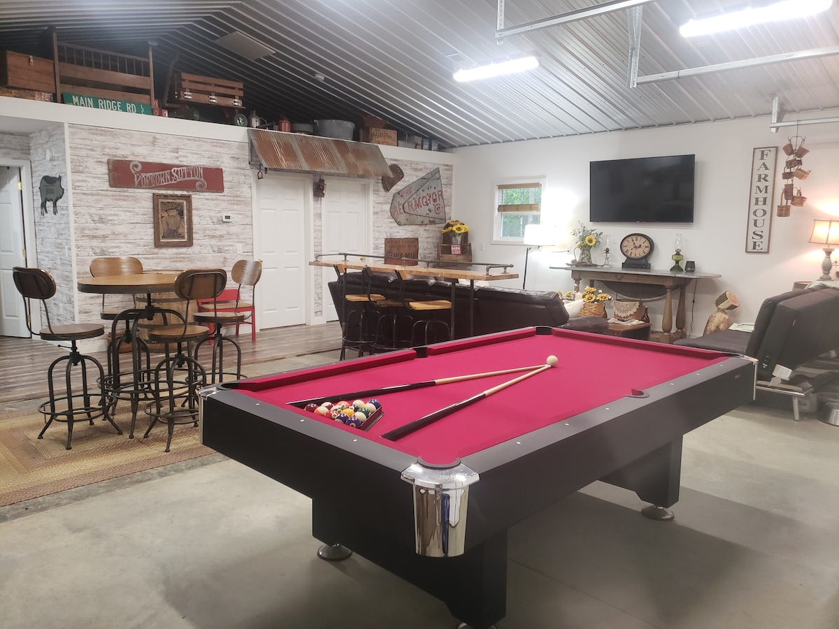 A game room features a red-felt pool table, with cue sticks resting on its side. In the background, a round high-top table and bar stools provide seating, while decorative elements like signs and a wall-mounted TV enhance the farmhouse theme.