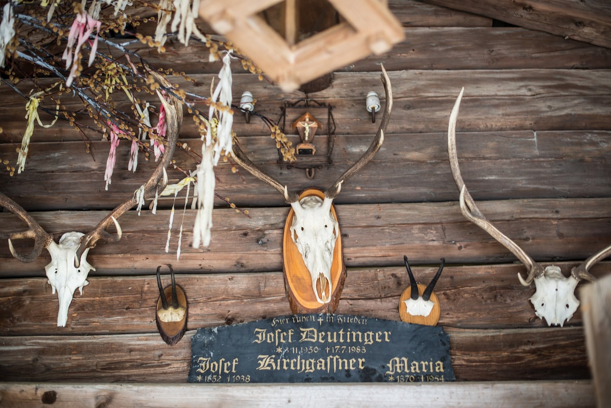 Mounted deer antlers and skulls are showcased on a rustic wooden wall. A decorative plaque with names is positioned centrally. Natural elements, such as dried flowers and branches, hang from above, contributing to the traditional alpine aesthetic.