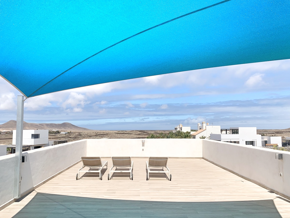 A spacious terrace features three lounge chairs under a large blue shade sail. Scenic views of distant hills and the surrounding landscape are visible against a partly cloudy sky.