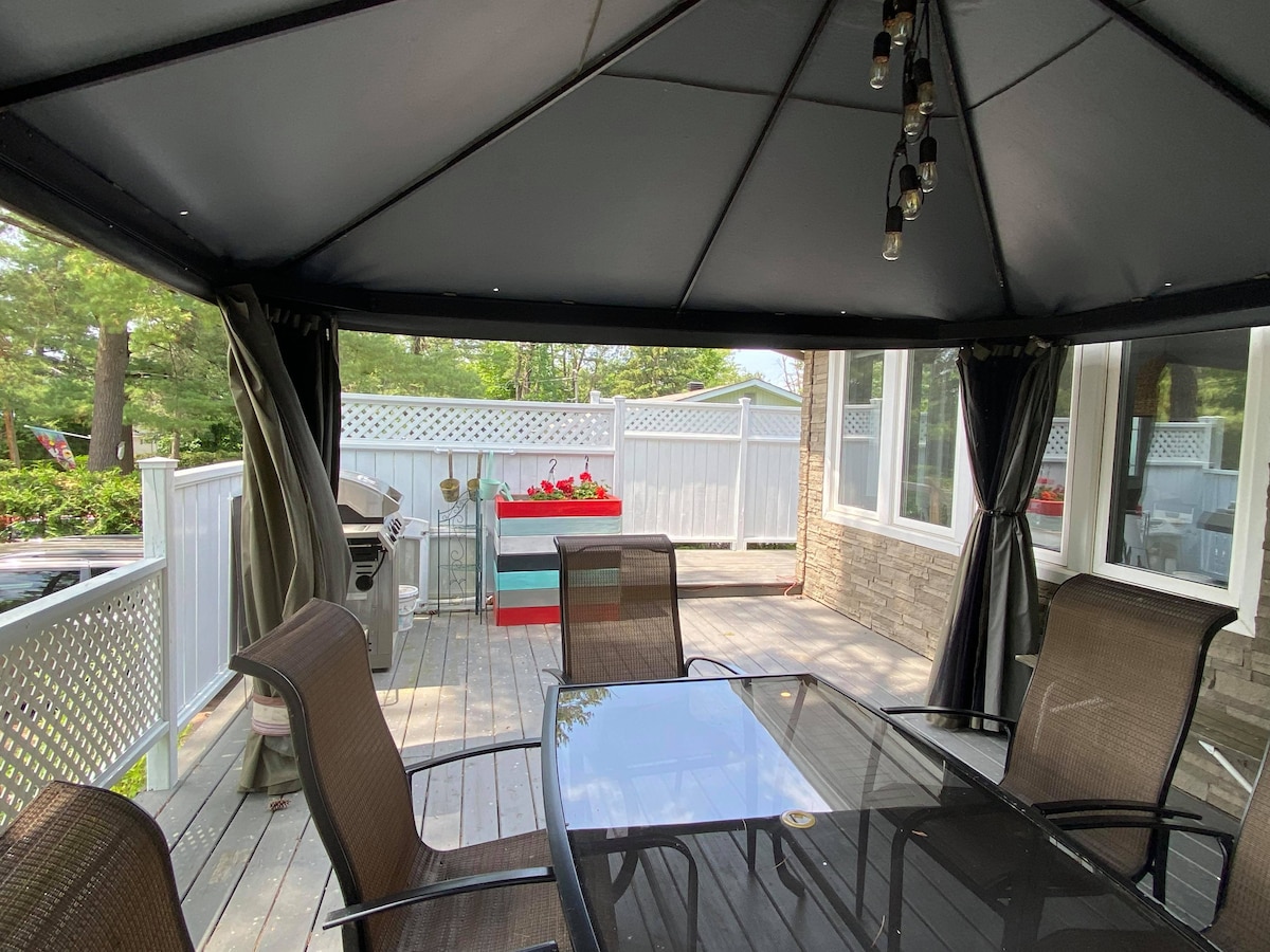 An outdoor gazebo is shown, featuring a glass-top table surrounded by six chairs. The space is enclosed by draped fabric and is illuminated by overhead lights. A propane BBQ grill is positioned nearby, with a backdrop of greenery and a white fence.
