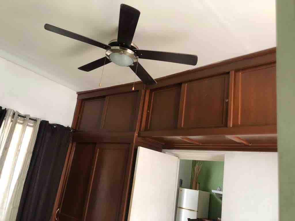A ceiling fan with dark wooden blades is mounted in the corner of a light-colored room. Wooden cabinetry is visible on the walls, providing ample storage space. Soft light enters through partially drawn dark curtains, creating a calm atmosphere. A white refrigerator is seen in the background.