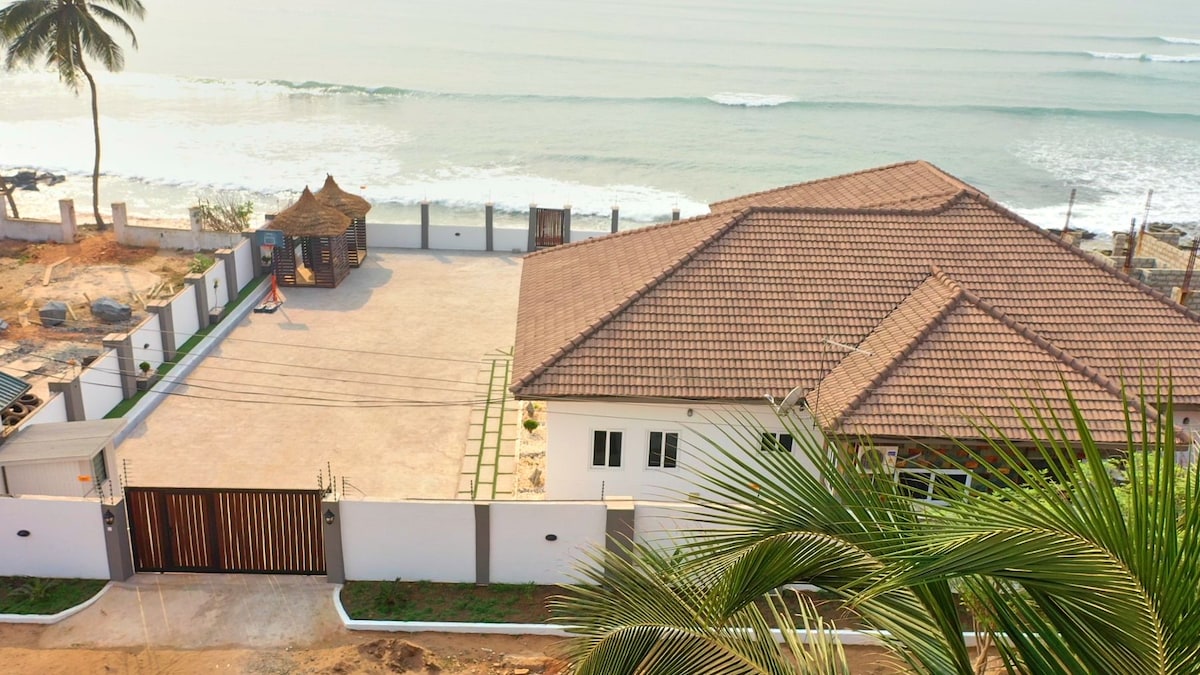 An aerial view captures a beachfront villa surrounded by gentle waves and greenery. The structured layout includes a spacious patio in front of the house, with a thatched cabana visible in the background, complementing the serene coastal environment.
