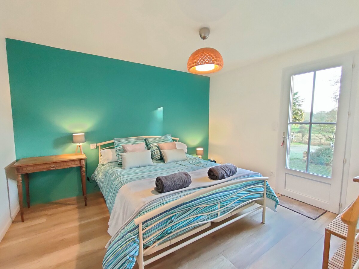 A spacious bedroom features a large bed adorned with striped bedding, complemented by two neatly placed towels. Soft lighting is provided by bedside lamps and a pendant light. A set of glass doors offers views of the surrounding greenery, enhancing the peaceful atmosphere.