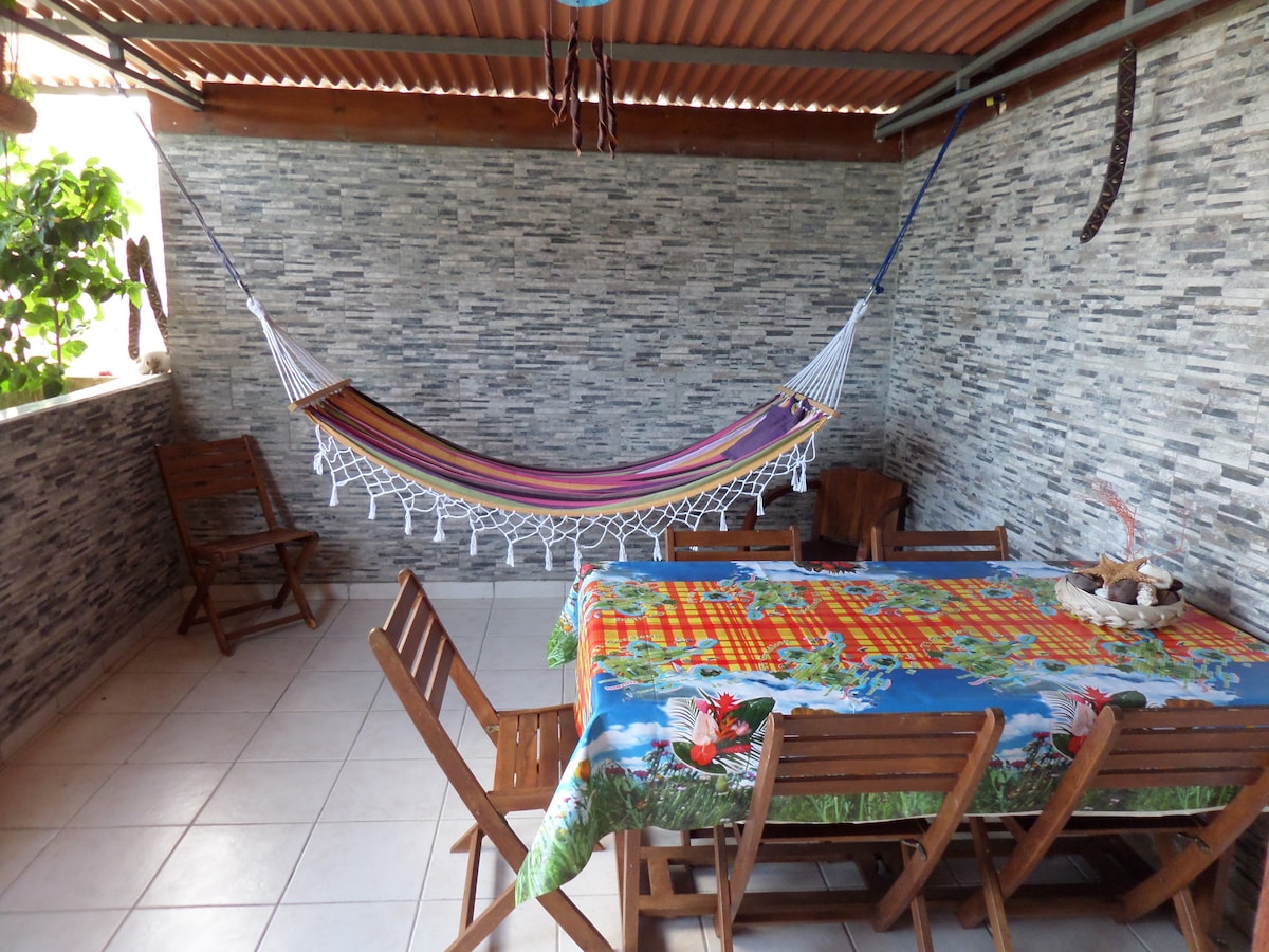 A shaded terrace features a vibrant table covered with a colorful tablecloth, surrounded by wooden chairs. A hammock hangs gracefully nearby, offering a spot for relaxation, while textured stone walls provide a rustic backdrop. Sunlight filters through, enhancing the outdoor ambiance.