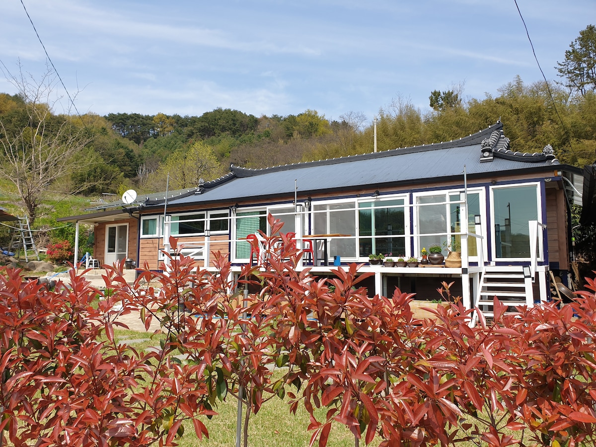 The traditional hanok is displayed with large glass windows and a welcoming porch, framed by vibrant red foliage. The building features a pitched roof with dark tiles and is surrounded by greenery, providing a serene rural setting.