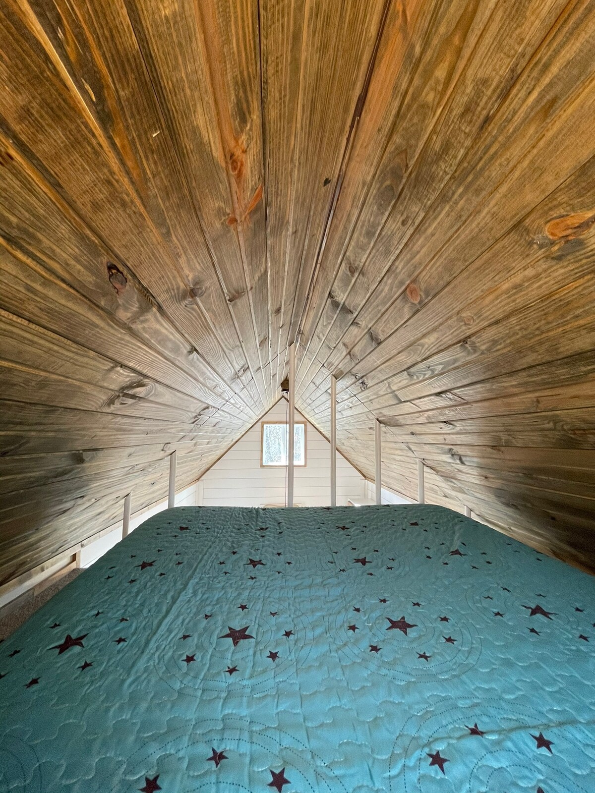 A lofted sleeping area features a spacious covered mattress topped with a star-patterned quilt. The wooden ceiling angles gracefully downward, and a small window allows natural light to illuminate the space. Warm wooden tones create a cozy and inviting environment.