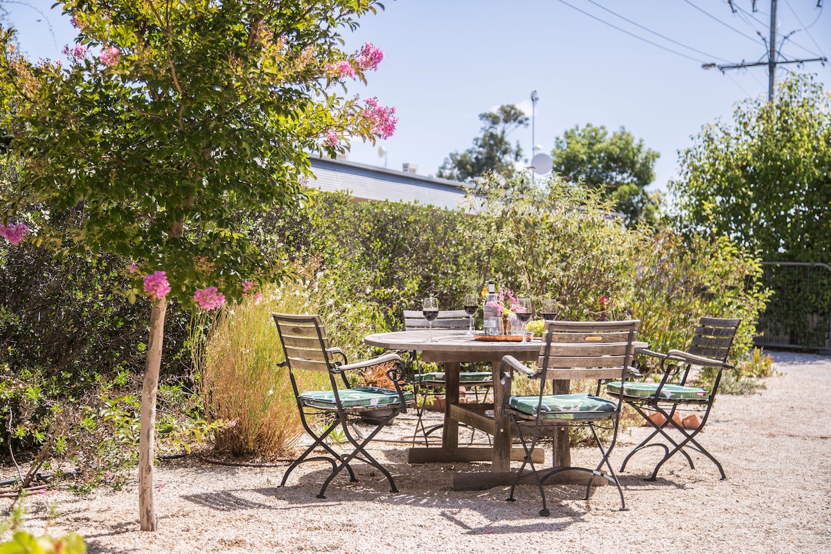 An outdoor patio area features a wooden table surrounded by several metal-framed chairs with cushions. Lush greenery, including flowering plants and shrubs, borders the space, creating a serene setting for relaxation and dining in the garden.