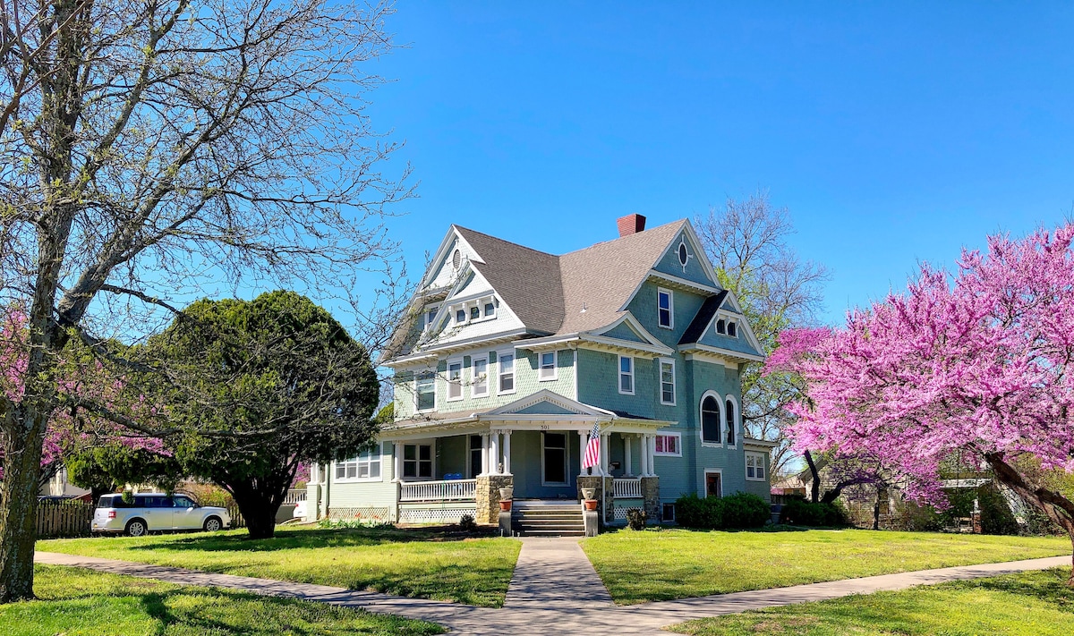 A historic mansion with a light blue exterior is framed by trees and vibrant pink blossoms. The wrap-around porch invites relaxation, while a well-kept lawn complements the home's elegant architecture. A white vehicle is parked nearby, providing a sense of accessibility.