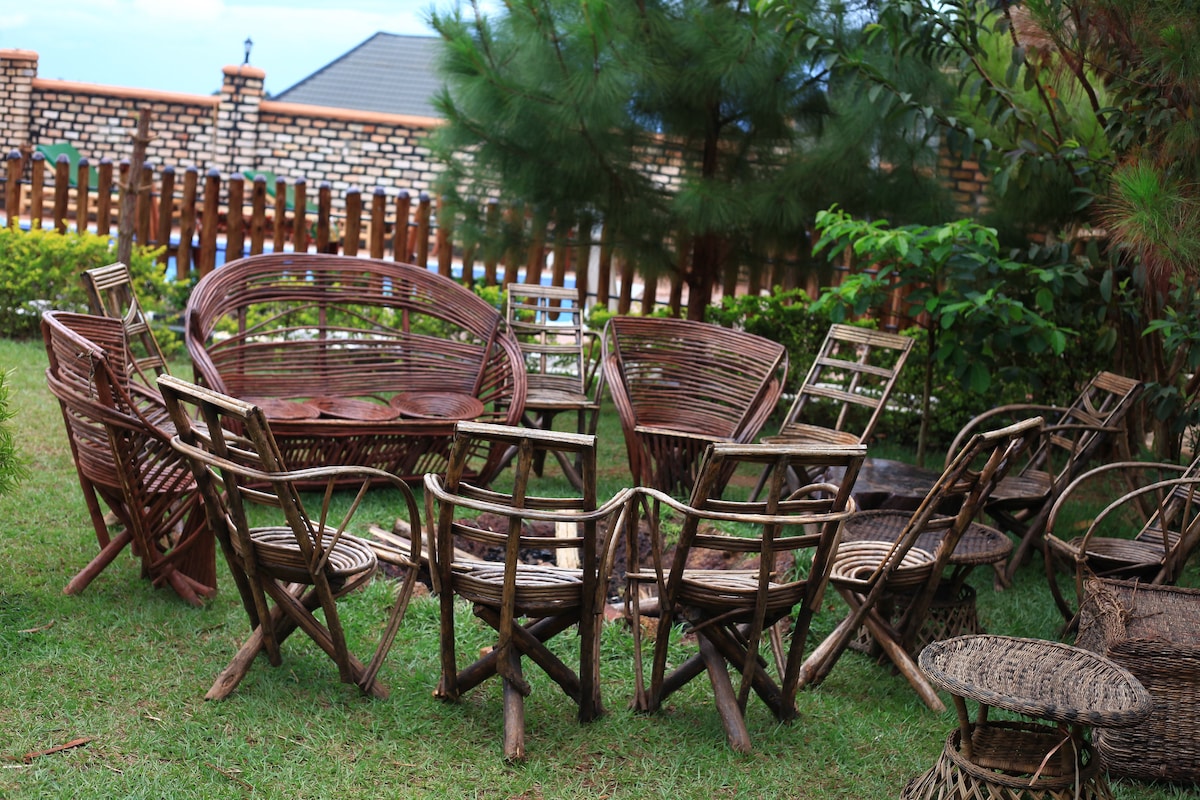 A collection of woven furniture is arranged in a lush outdoor setting. Various styles of chairs and tables, crafted from natural materials, are surrounded by greenery. A wooden fence is visible in the background, providing a sense of privacy to the space.
