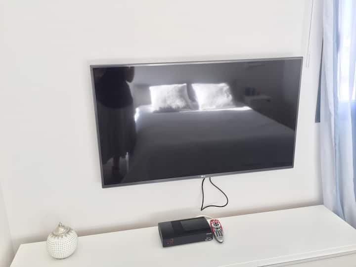 Chumba kikuu cha kulala kina televisheni mahiri na kebo ya inchi 42. Tafakari ya kitanda cha ukubwa wa malkia kwenye televisheni. 