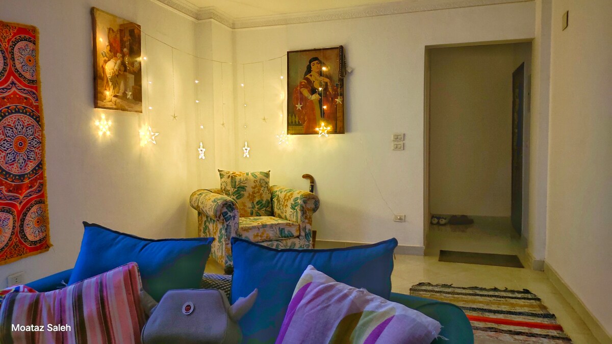 A cozy living area features a vibrant blue couch adorned with colorful throw pillows. A patterned rug lies underneath, while a floral armchair is positioned nearby. Decorative wall art and twinkling string lights create a relaxed, inviting ambiance.