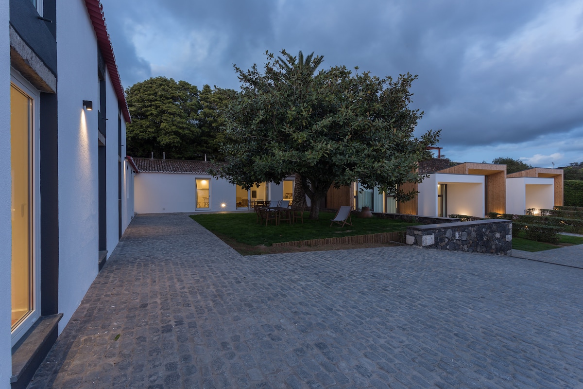 The outdoor area showcases a cobblestone path leading to the property, framed by a large tree and soft garden lighting. The evening sky is filled with clouds, while the beautifully designed guest house features warm-lit windows, hinting at inviting spaces within.