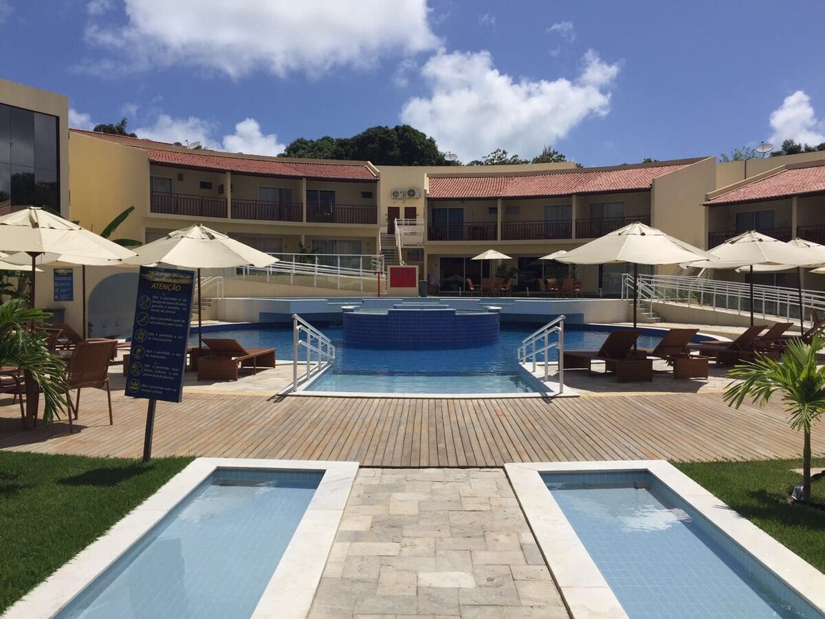 A well-maintained outdoor area features a central swimming pool surrounded by wooden decking. Lounge chairs with umbrellas provide shaded seating options. Clear pathways lined with greenery lead to individual units, while additional pools are visible in the distance under a blue sky.
