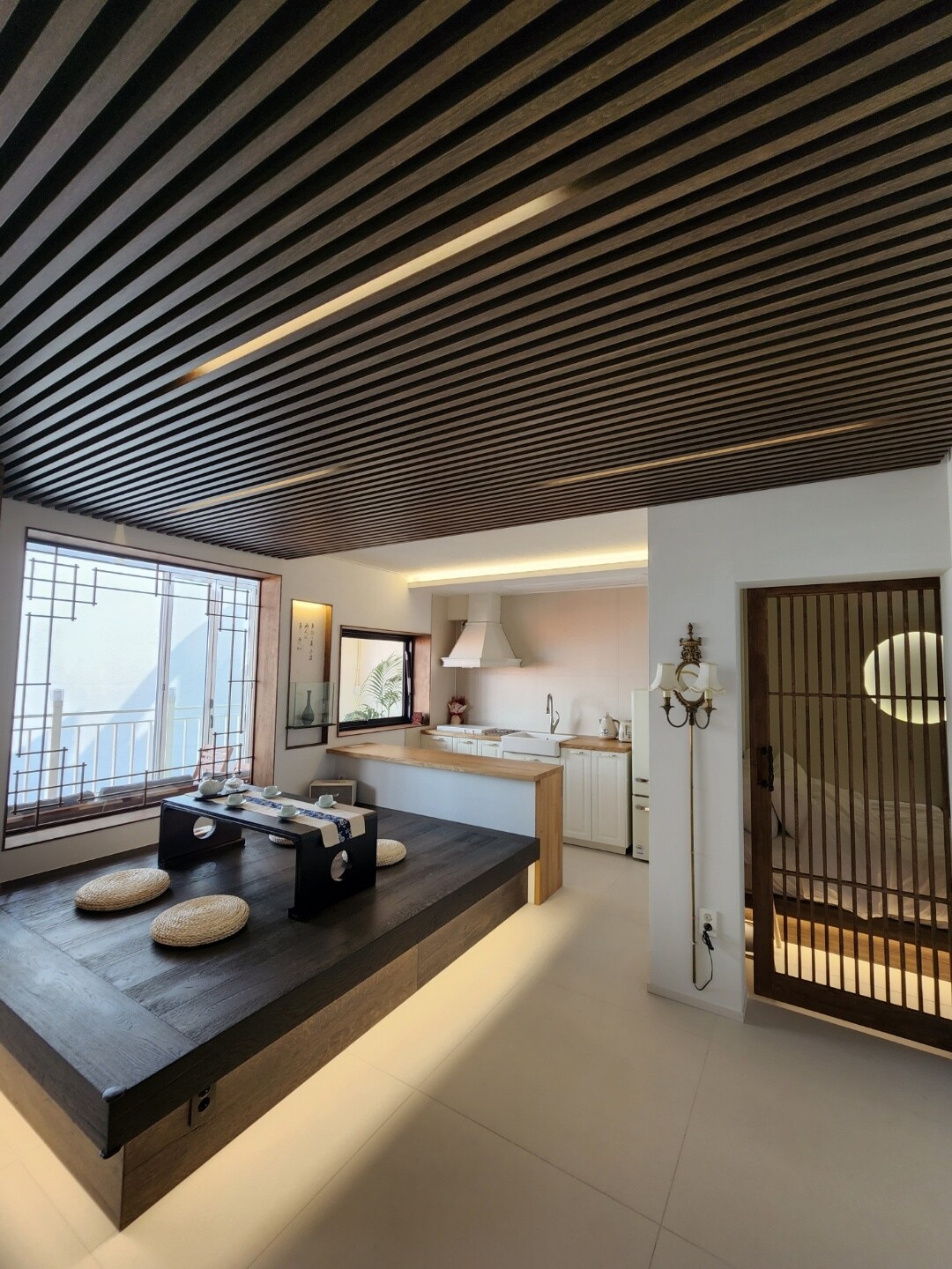 A modern kitchen space is visible, featuring a minimalist design with wooden beams on the ceiling. A low seating area with cushions is positioned beside large windows, allowing natural light to fill the room. The kitchen appliances are neatly integrated, creating an open and inviting layout.