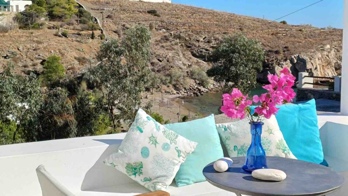 A serene terrace area is highlighted by a small round table, adorned with a blue glass vase holding vibrant pink flowers. Soft cushions in soothing turquoise and light blue tones are arranged on a white seating area, creating a peaceful space overlooking a natural landscape.