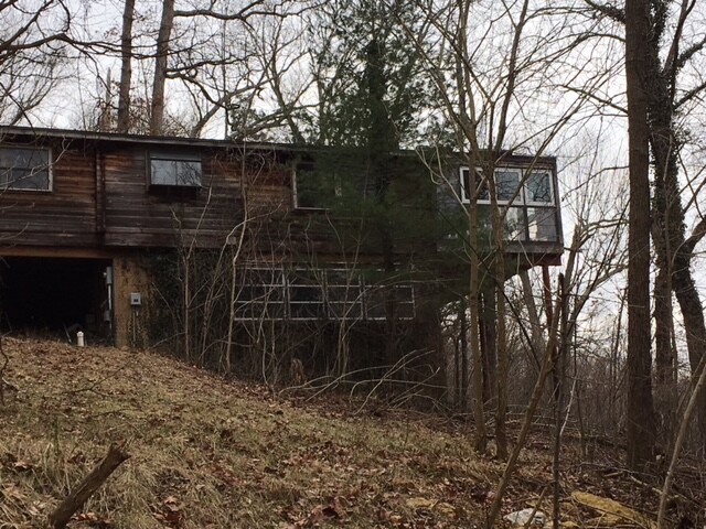 A two-story cabin is nestled among trees, featuring wooden siding and large windows that provide natural light. The structure is elevated slightly from the ground, with a sloped, grassy area in the foreground. Surrounding foliage includes bare branches and evergreen trees.