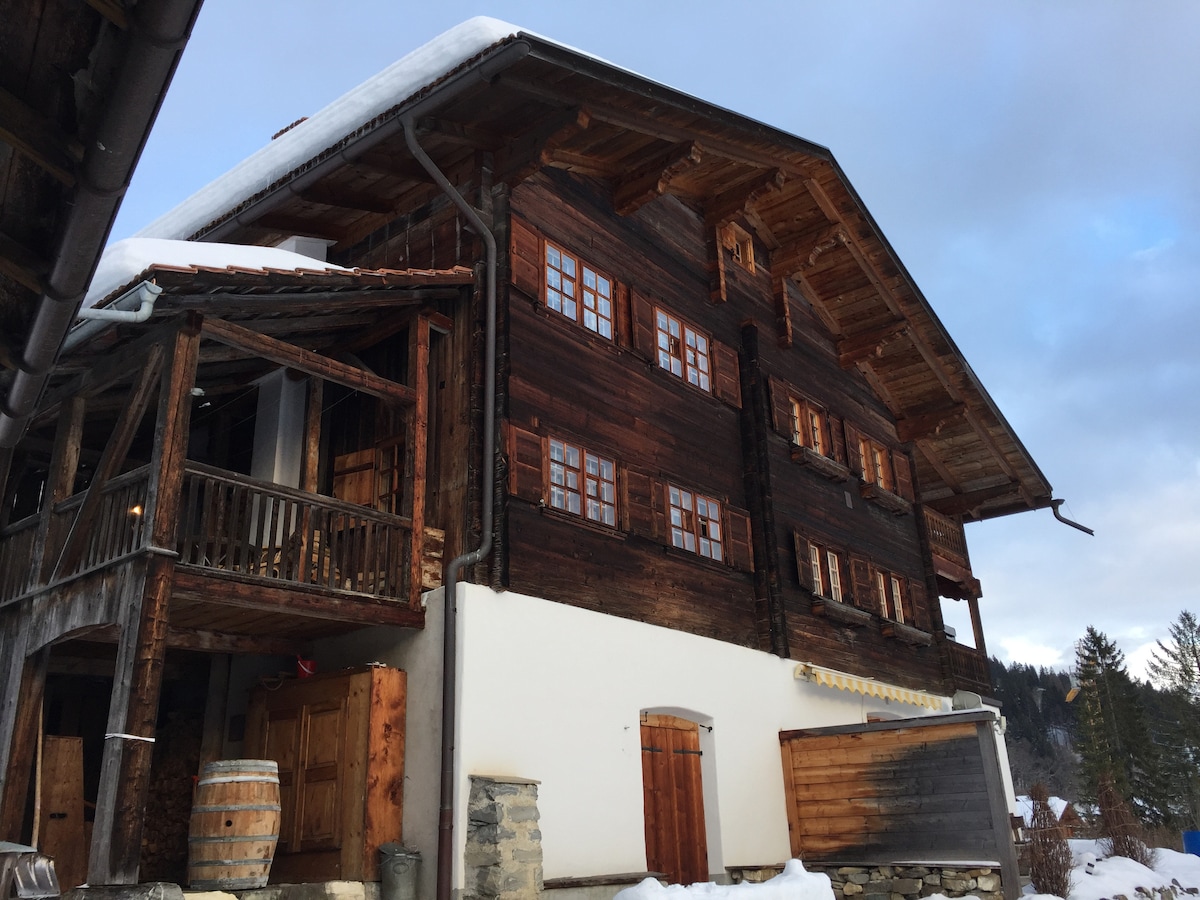The exterior of a charming wooden chalet is displayed, featuring large, multiple-pane windows and a sloped roof. Snow covers the ground, while wooden barrels and traditional doors are visible. The structure exemplifies rustic alpine architecture, set against a backdrop of trees and a cloudy sky.