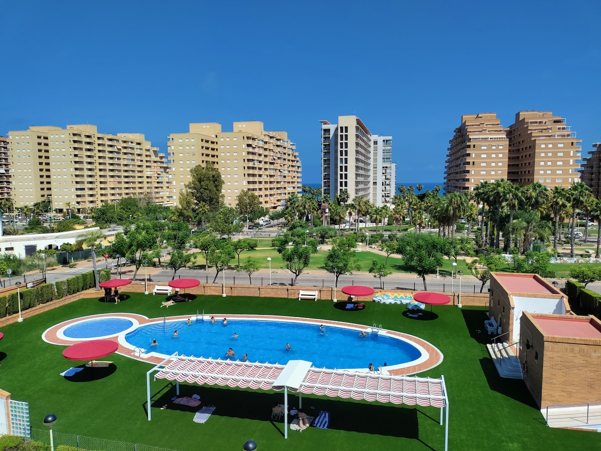 A large swimming pool is surrounded by well-maintained green lawn and palm trees. Red umbrellas provide shade on the pool deck. In the background, modern buildings rise against a clear blue sky, with hints of the ocean visible in the distance.