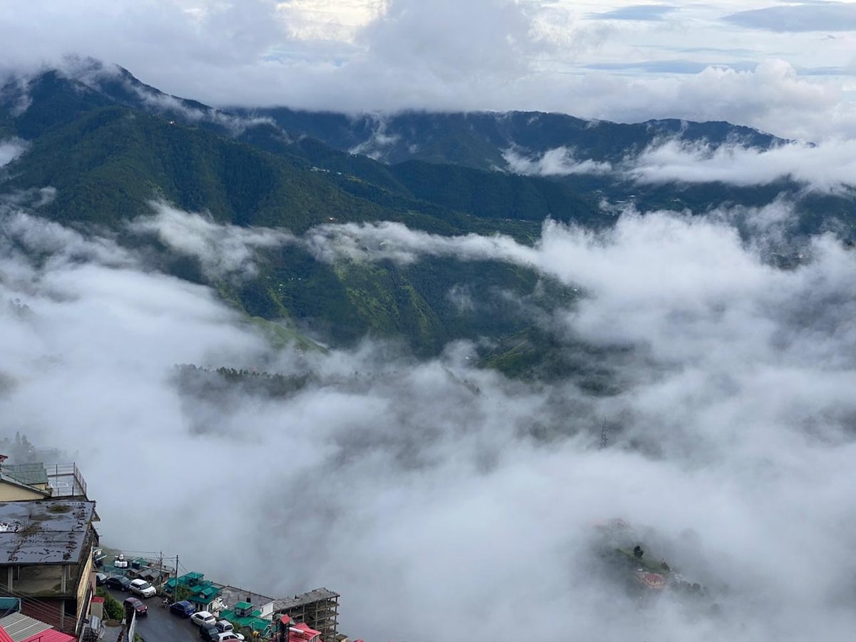 A panoramic view of mist-covered mountains is presented, with layers of clouds enveloping the lower valleys. Brightly colored buildings can be glimpsed at the image's base, while the peaks are partially obscured by the fog.