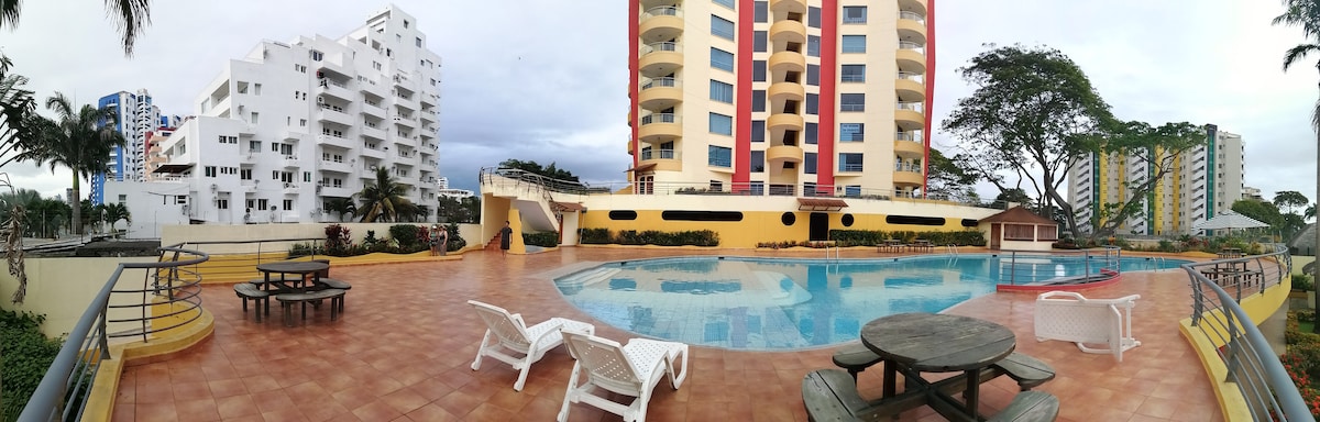 A panoramic view of the pool area reveals a rectangular swimming pool surrounded by tiled decking. Sun loungers are positioned near the water, and a shaded seating area is visible in the foreground. The multi-story building provides a backdrop, framed by lush greenery.