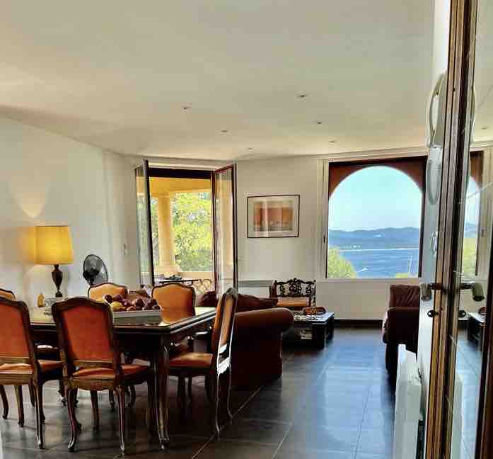 A spacious living area is presented with large, open glass doors that invite natural light and offer views of the bay. A dining table sits surrounded by elegant chairs, while a comfortable sofa adds to the welcoming environment. Subtle decor elements enhance the calm atmosphere.