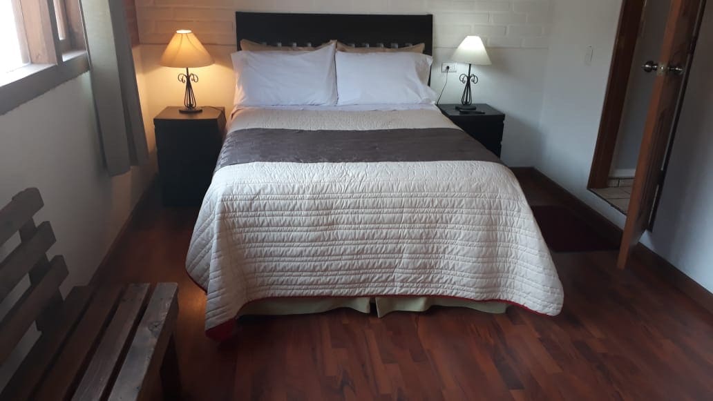 A cozy sleeping area features a spacious bed covered with a light quilt. Nightstands with lamps are positioned on either side of the bed, providing subtle illumination. Warm wood flooring enhances the inviting atmosphere, while a wooden bench adds functional seating near the door.