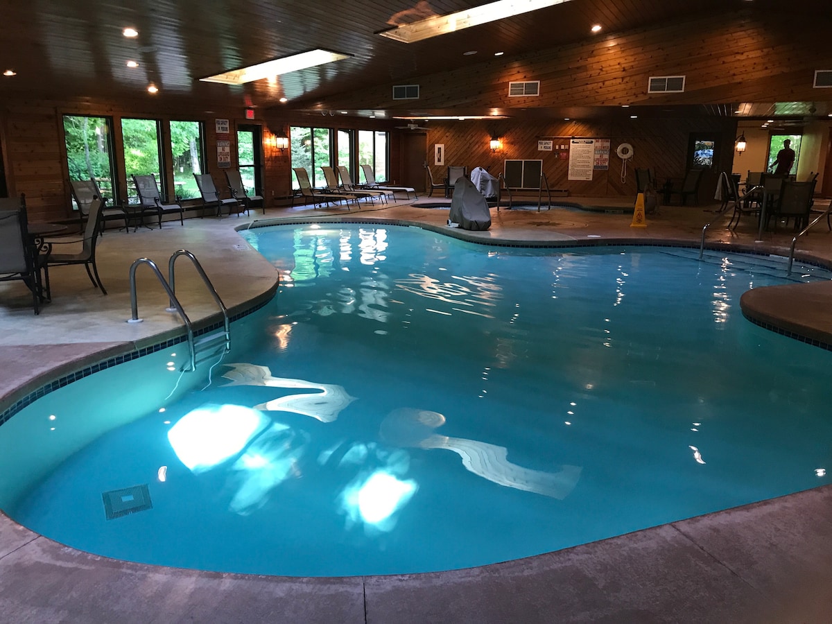 An indoor pool area is surrounded by wooden walls and large windows that allow natural light to fill the space. Several lounge chairs are positioned around the water, which is clear and illuminated, creating a serene atmosphere.